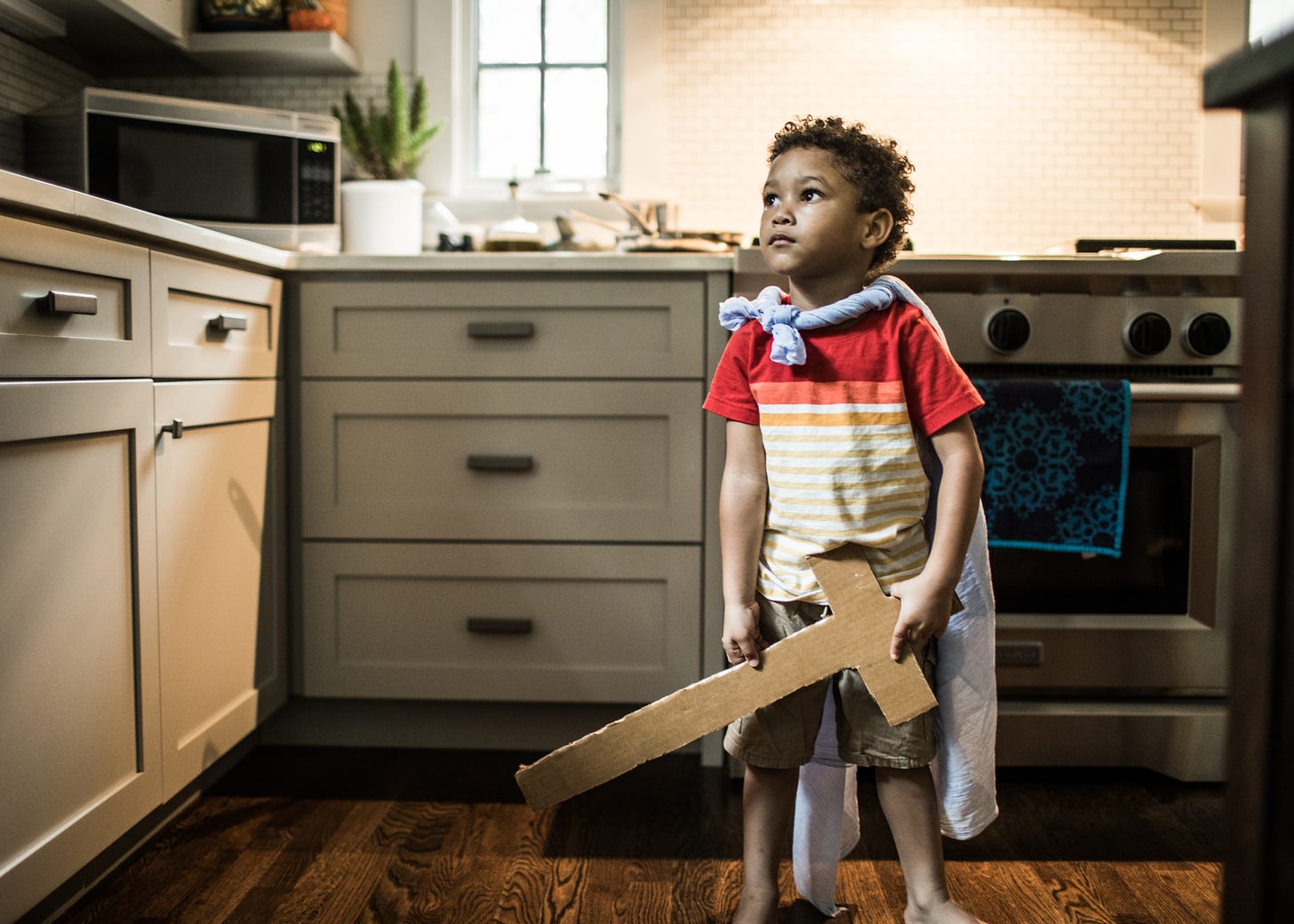 A child in a kitchen with a cardboard sword and homemade cape