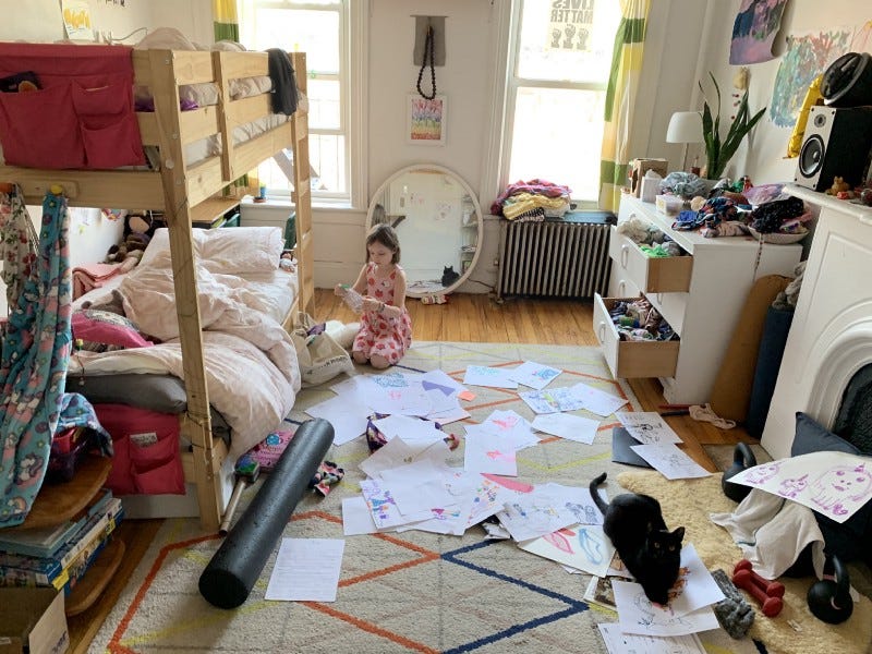 a seven year old child sits in her room amidst a bunch of papers on the ground. a black cat is in the foreground and bunk beds are on the left. she is reading.