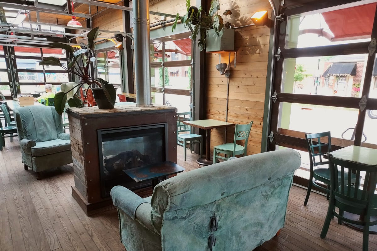The inside of a coffee shops seating area. A faux fireplace with a silver exhaust column sits in the middle of the room and middle of the frame. It opens to both sides. Worn and tattered denim covered lounge chairs face the fireplace and rolling garage doors line the wall. Low slung tables with chairs are pushed against the garage door wall, and house plants are mounted on pine wood walls between the doors.