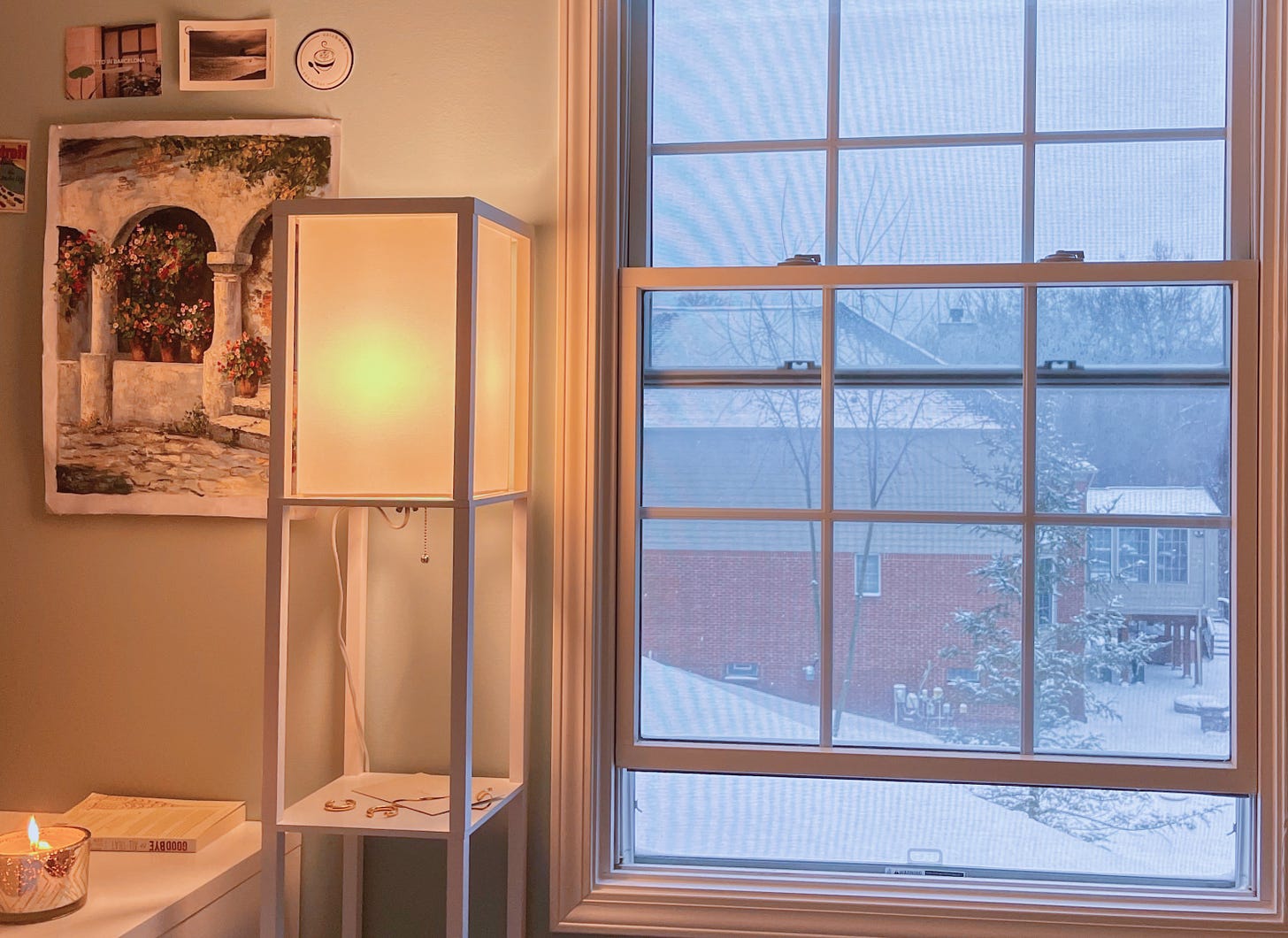 a snowy window, a white lamp next to a desk with a candle and a painting of a stone arch above  it