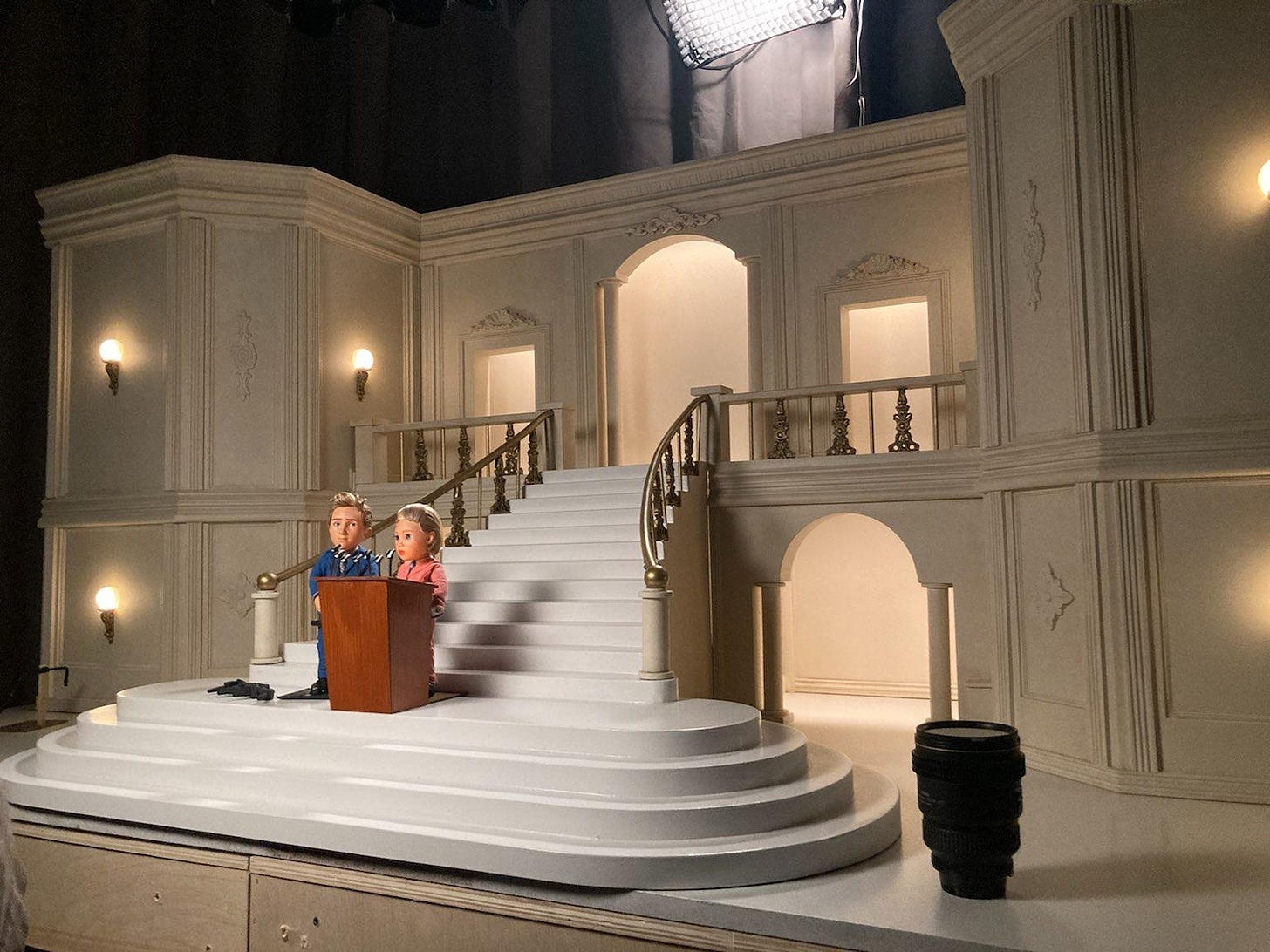 2 puppets stand at a podium, super prep and poised politician. They are at a podium infront of this grand white miniature building that is the mayors office. A lens is on the table to scale