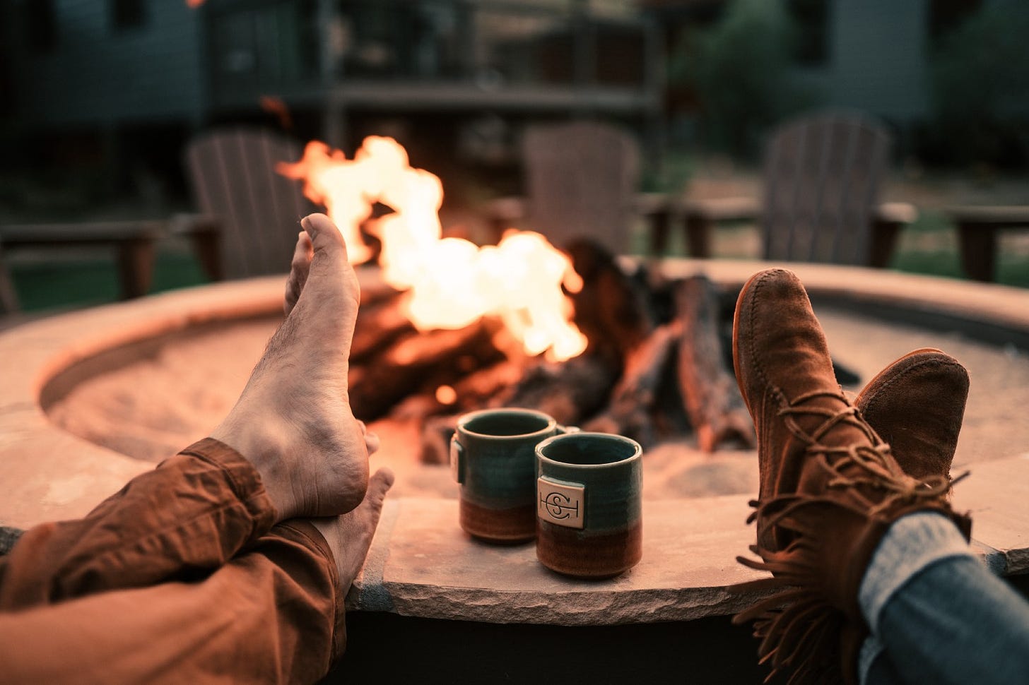 Two sets of feet rest on the edge of a large fire pit. Mugs sit between the feet. A fire burns in the background.