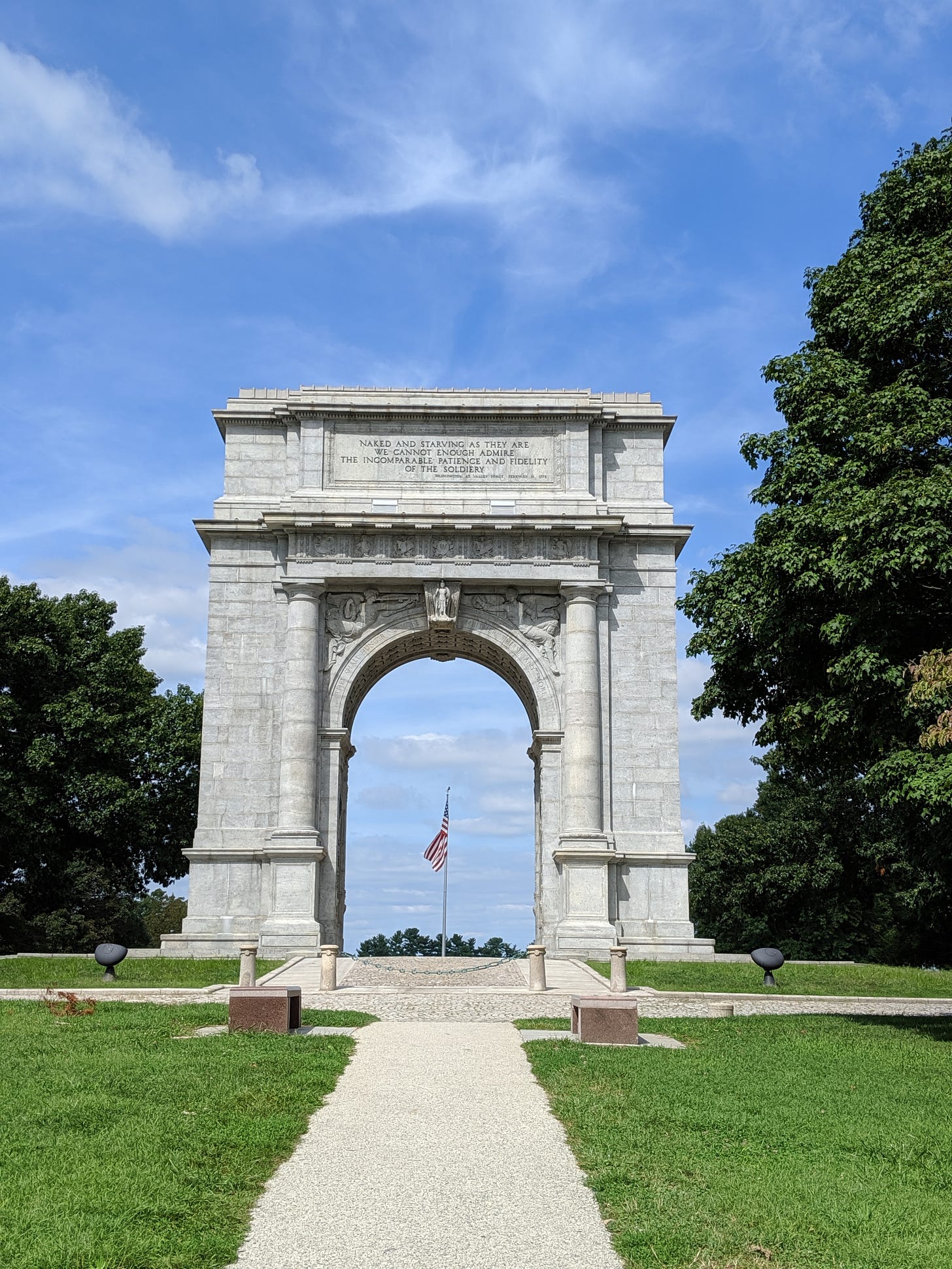National Monument Arch at Valley Forge. A large stone arch rises against a blue sky. An American flag is visible through the arch.