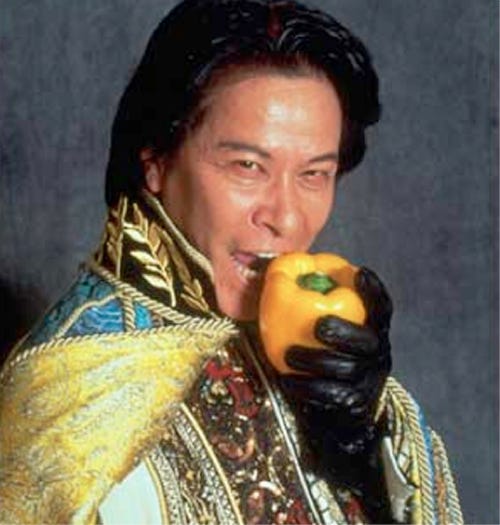 Iron Chef Chairman Takeshi Kaga bites in a pepper while starring at the camera. He's in his best cape. 
