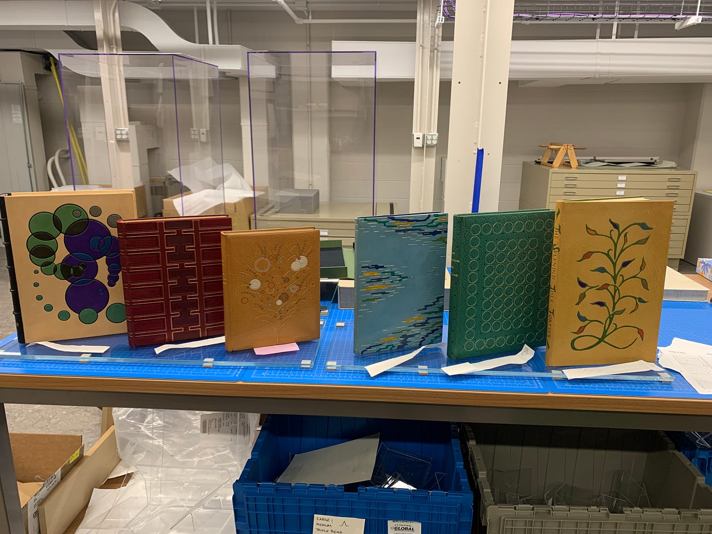 Image of six fine bindings standing upright on a worktable. The binding of note is on the far right, in tan leather, with a looping bean stalk design in green to the center of the upper cover. The leaves are in multiple colors: red, purple, yellow, green, blue, and orange, like tiny leather jewels.