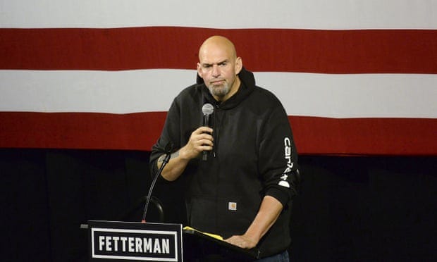 Man wearing a hoodie with rolled up sleeves standing behind a podium, with a mike in one hand and the American flag in the background