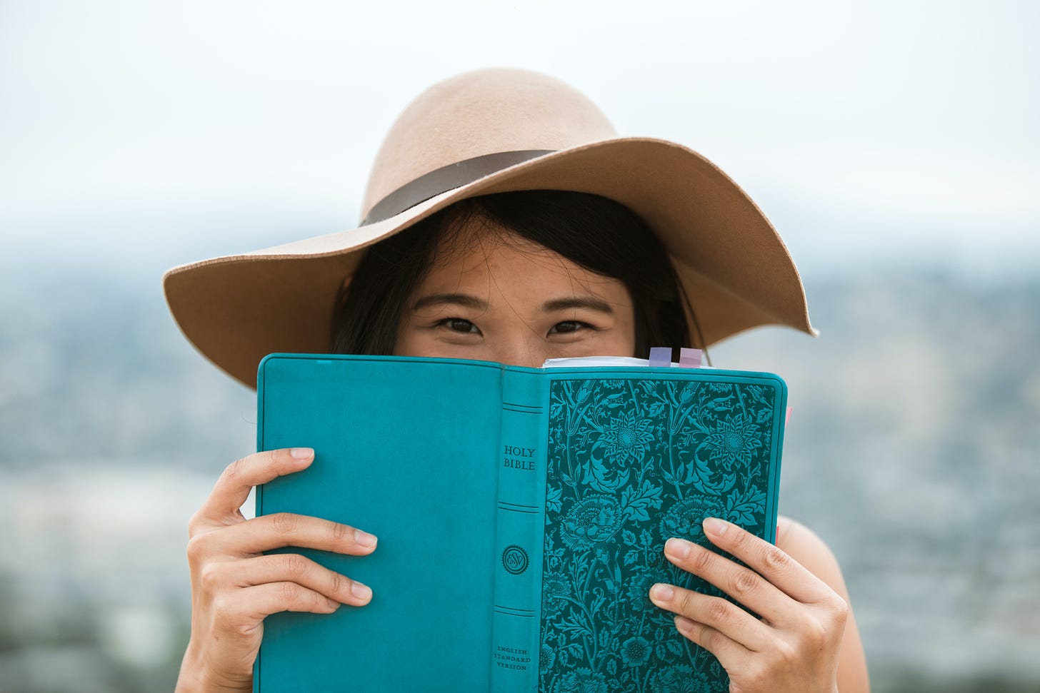 An Asian woman wearing a hat looking over the top of a blue Bible.