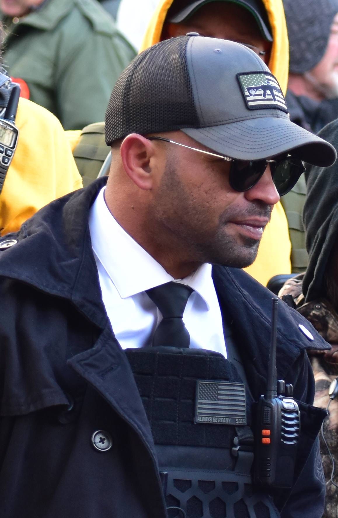 A picture of Enrique Tarrio in sunglasses and a suit, sitting in a crowd. He is wearing body armor under his suit jacket and a very official-looking communications device is strapped to it