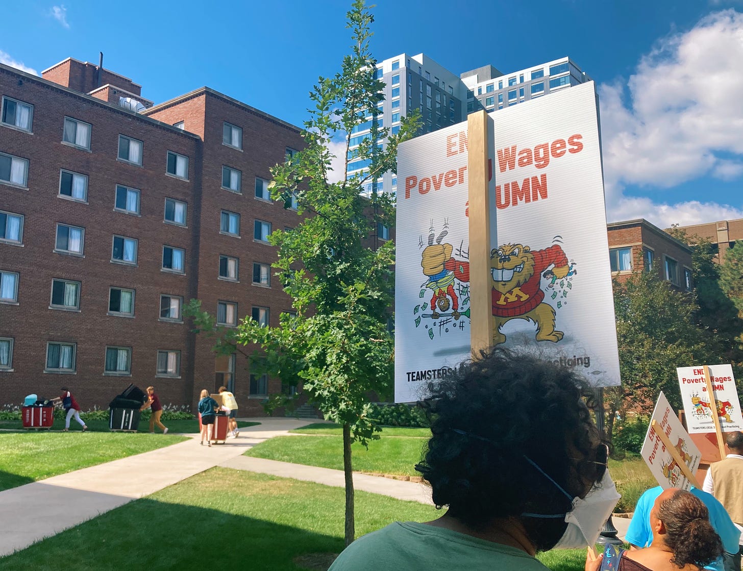 a man with curly black hair holds a sign reading "end poverty wages at UMN" while students moving in to campus push carts of belongings to the dorm buildings