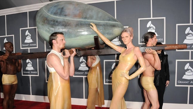 Photo of Lady Gaga arriving to the 2011 Grammys red carpet. Four performers carry a translucent blue egg with Gaga inside. Another performer poses outside the egg. This is the hardest alt text I've ever written.