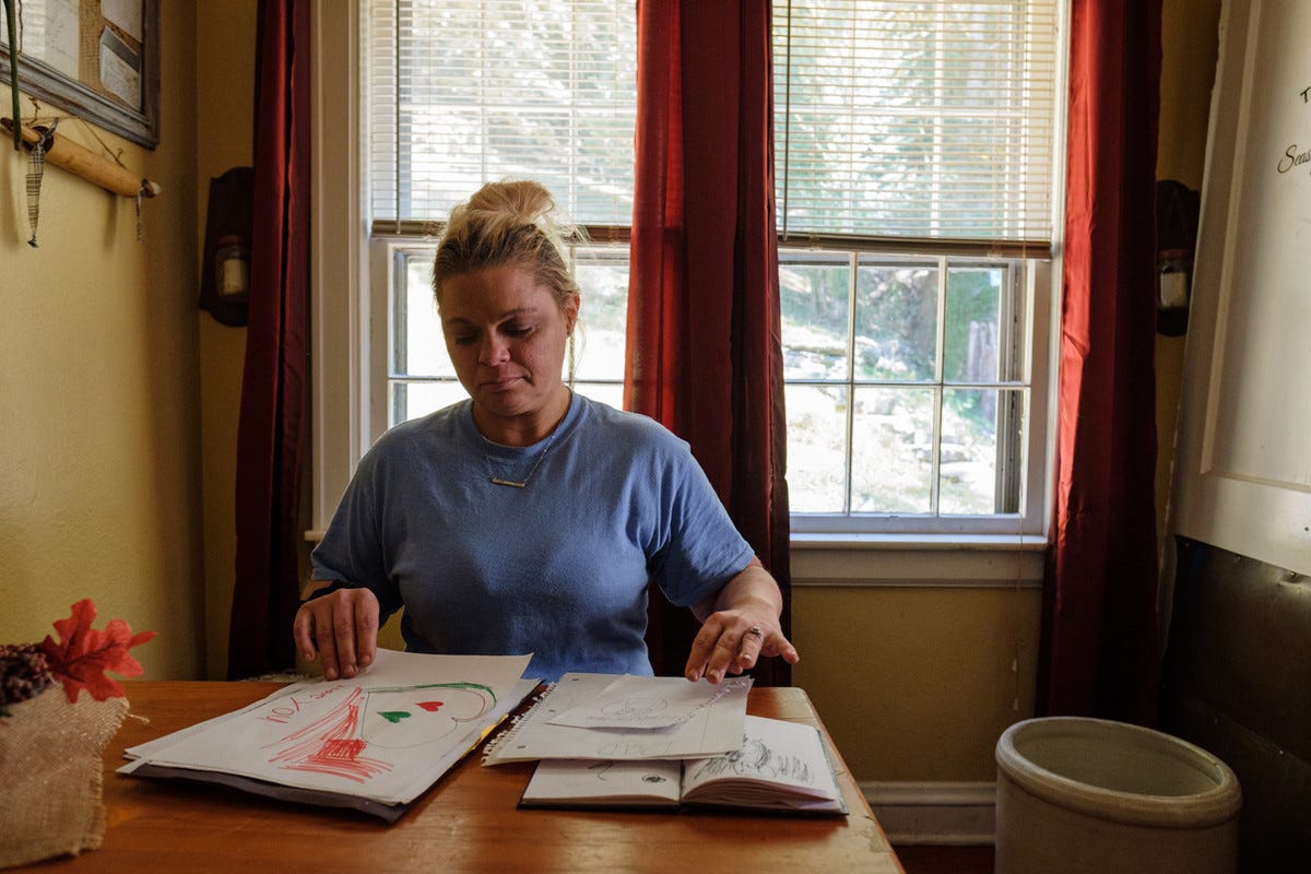 Jackie Snodgrass, a white woman with blonde hair pulled back into a messy bun and wearing a light blue top, sits at a table and looks down at her children’s drawings spread across the surface.