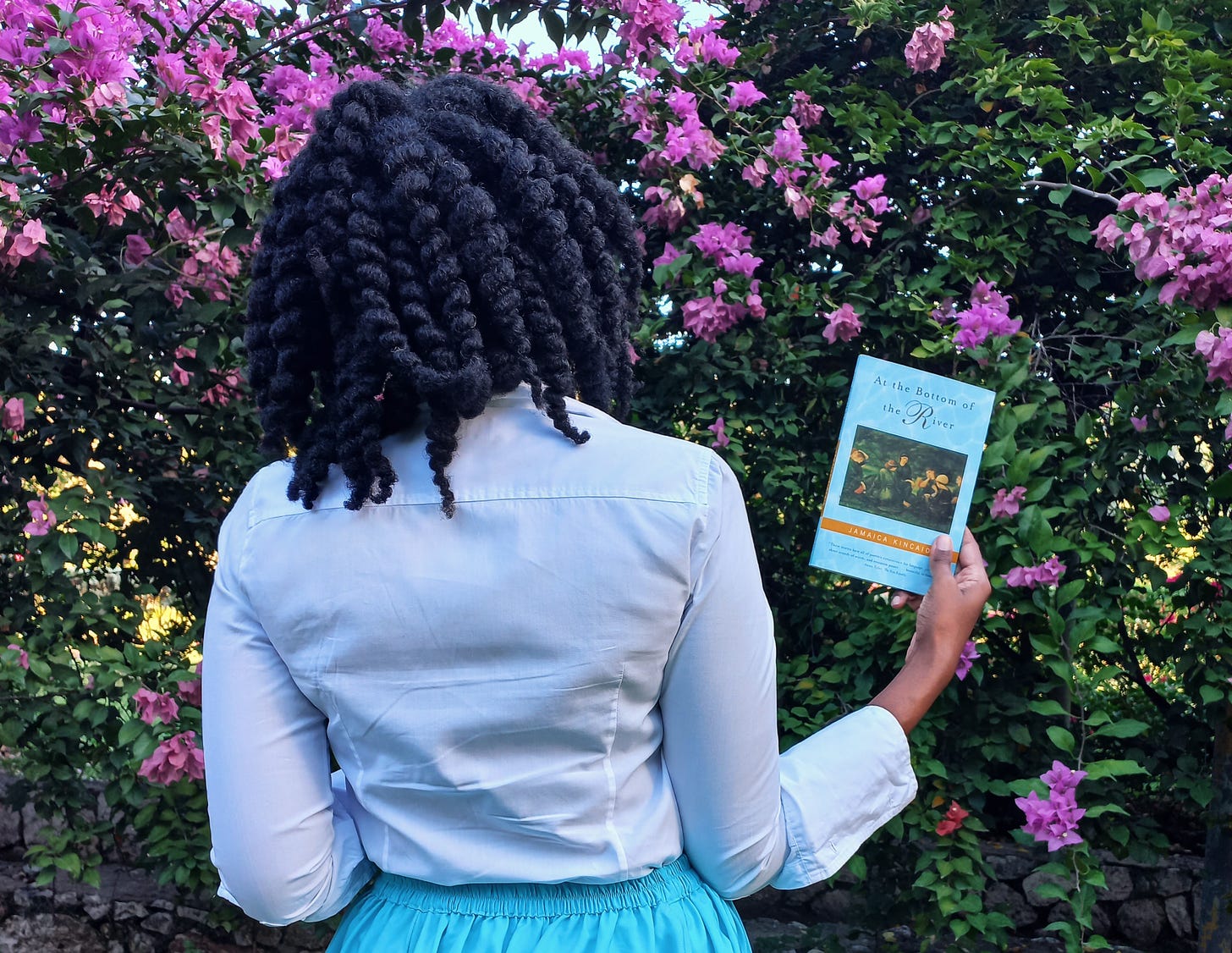 A black woman has her back to the camera holding a copy of the book visible to the viewer. She has thick natural hair in a loose twist out and wears a long white sleeve shirt and long blue skirt. only from the waist up is visible. she stands in front of an arch pink bougainvilleas.