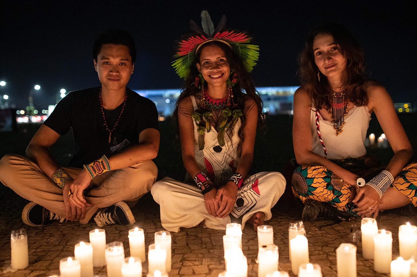 A young man, an indigenous woman and a European woman sit in front of candles and smile at the camera. It is night-time.