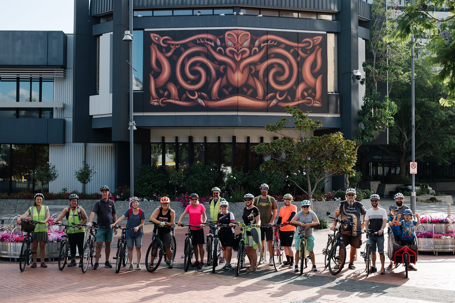 A photograph of a group of cyclists, standing outside the Hamilton City Council chambers. On the side of the chambers, behind the cyclists, is a large mural that looks like a Maaori wood carving. In the middle is the depiction of a tuupuna, with taniwha on each side.