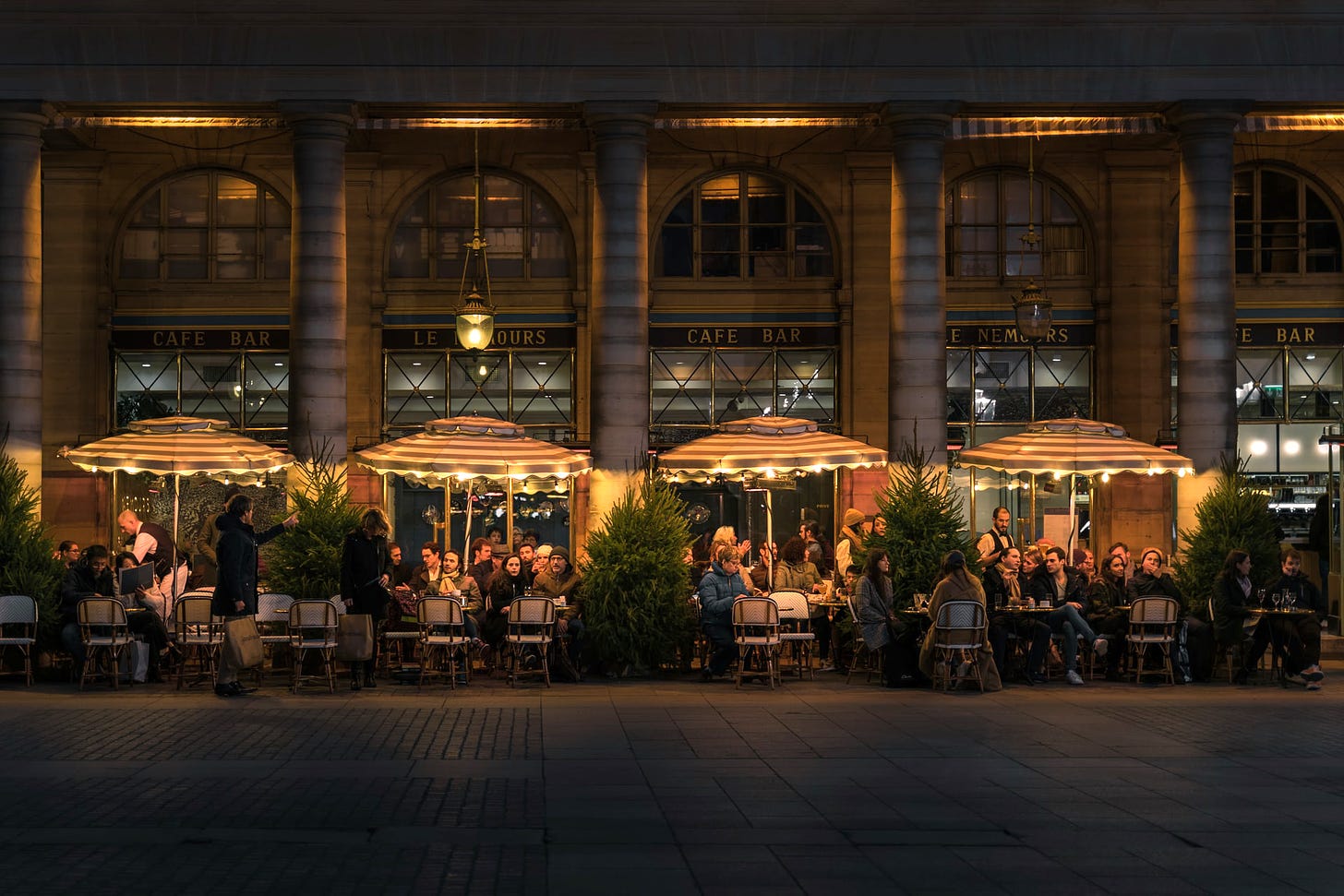 A photo of a Parisian street cafe at night. Many people are seated outside, under illuminated umbrellas.