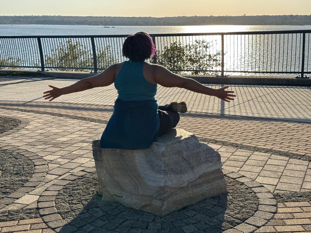 Kristen, a person with purple kinky hair, sits cross-legged on a rock, arms outstreched, facing a riverfront and the sun.