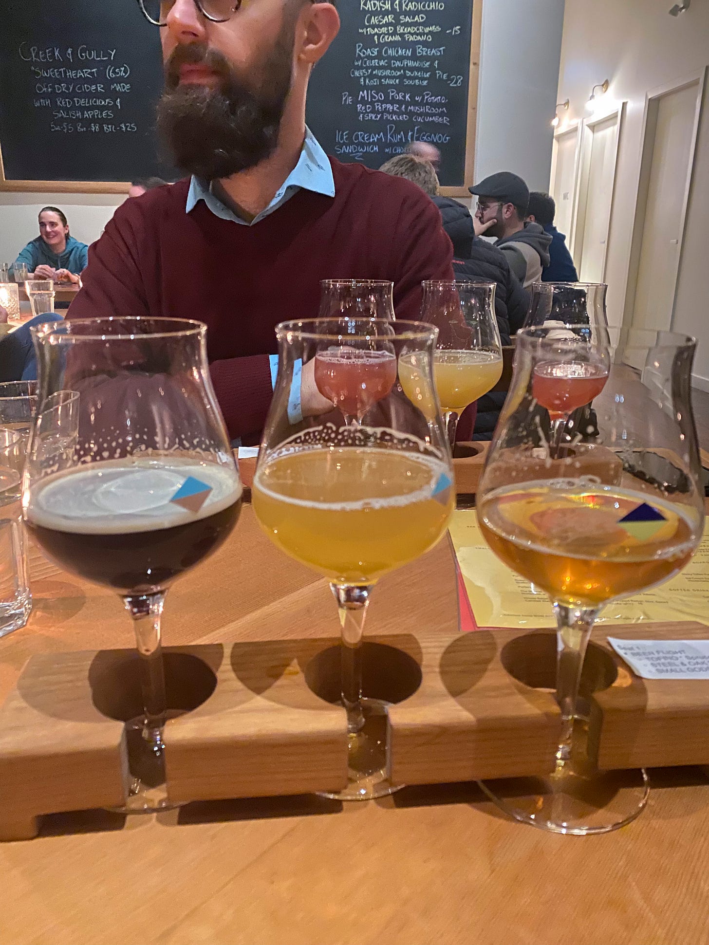 A flight of three beers in tulip glasses in the foreground, ranging from light to dark. Behind it is another flight, sitting in front of Jeff, whose face is partially visible at the top of the frame. In the background is another table of people, and a blackboard of specials on the wall.