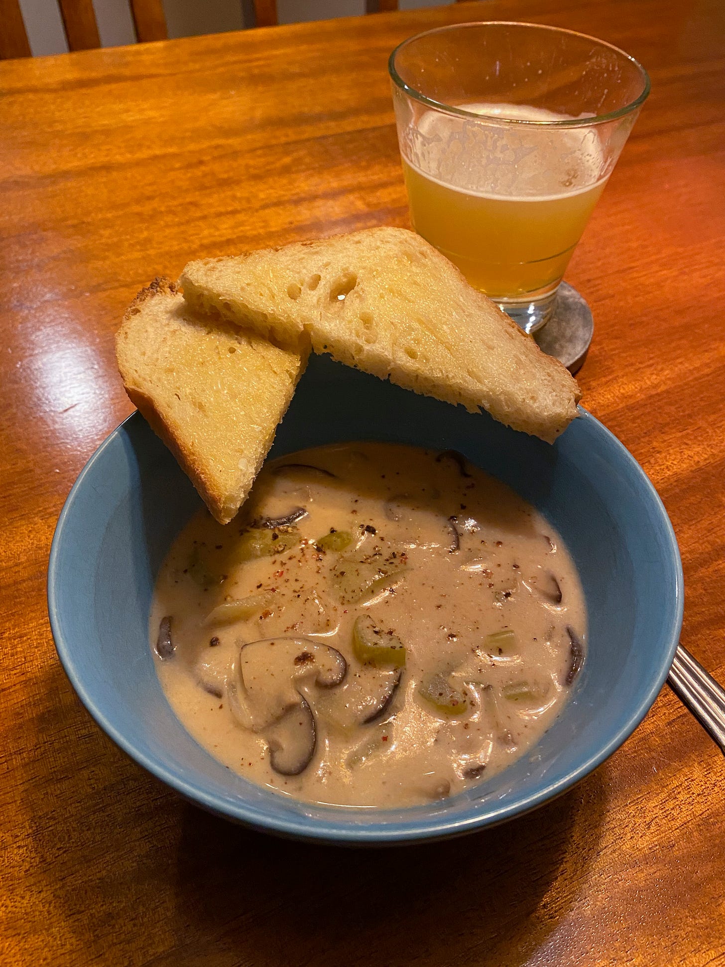 A blue bowl with vegan chowder, full of shiitake mushrooms, celery, and potatoes in a creamy base. Two triangles of toast rest on the side of the plate. A glass of IPA is on a coaster beside it.