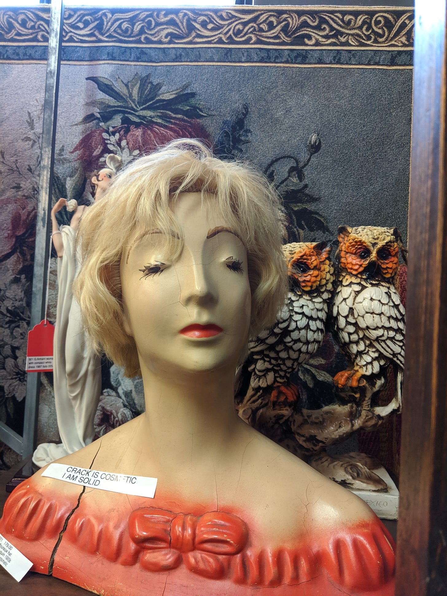 Photograph: a vintage mannequin head with closed eyes, creepy long eyelashes and blond hair (a wig?); figure is cracked, and a note on the figure says "CRACK IS COSMETIC / I AM SOLID"