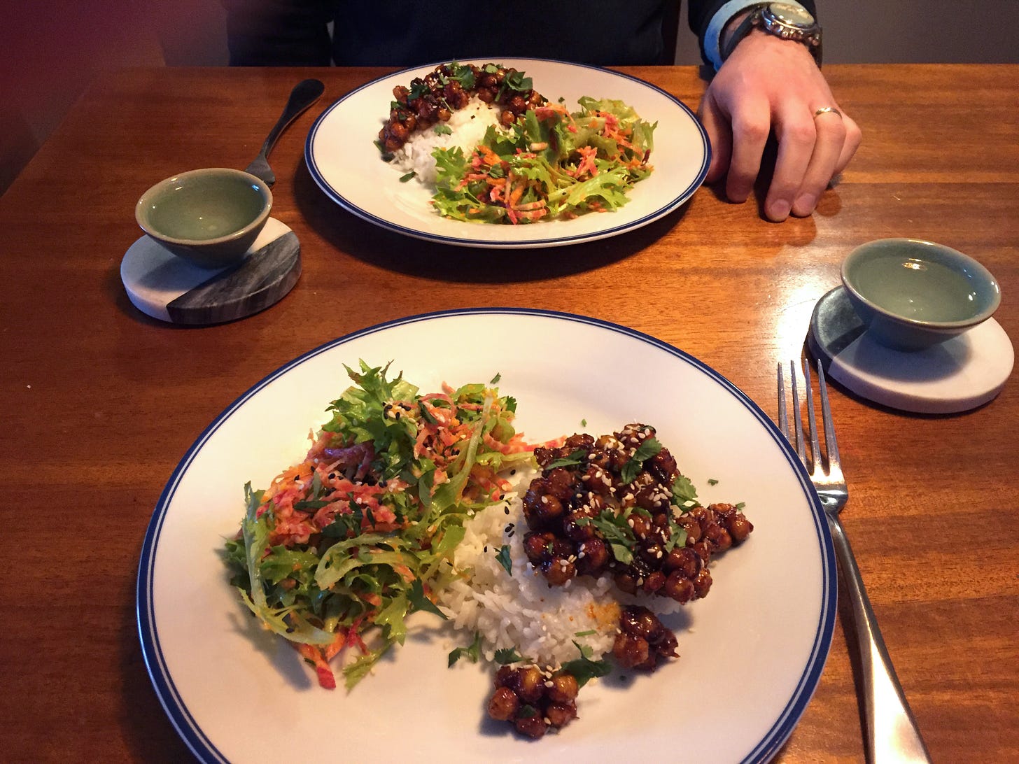 Two plates across from each other on the table, each about half full of a green salad with shredded Chiogga beet and carrot, and the other half filled with white rice and dark red glazed chickpeas. Cilantro and sesame seeds are sprinkled on top. Two small cups of soju sit on coasters next to the plates.