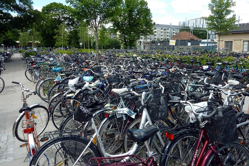A lot of bicycle parking at a railway station in Sweden. There is a line of trees in the background. There are a lot of bicycles. too many to count. Of all sorts of shapes and sizes.