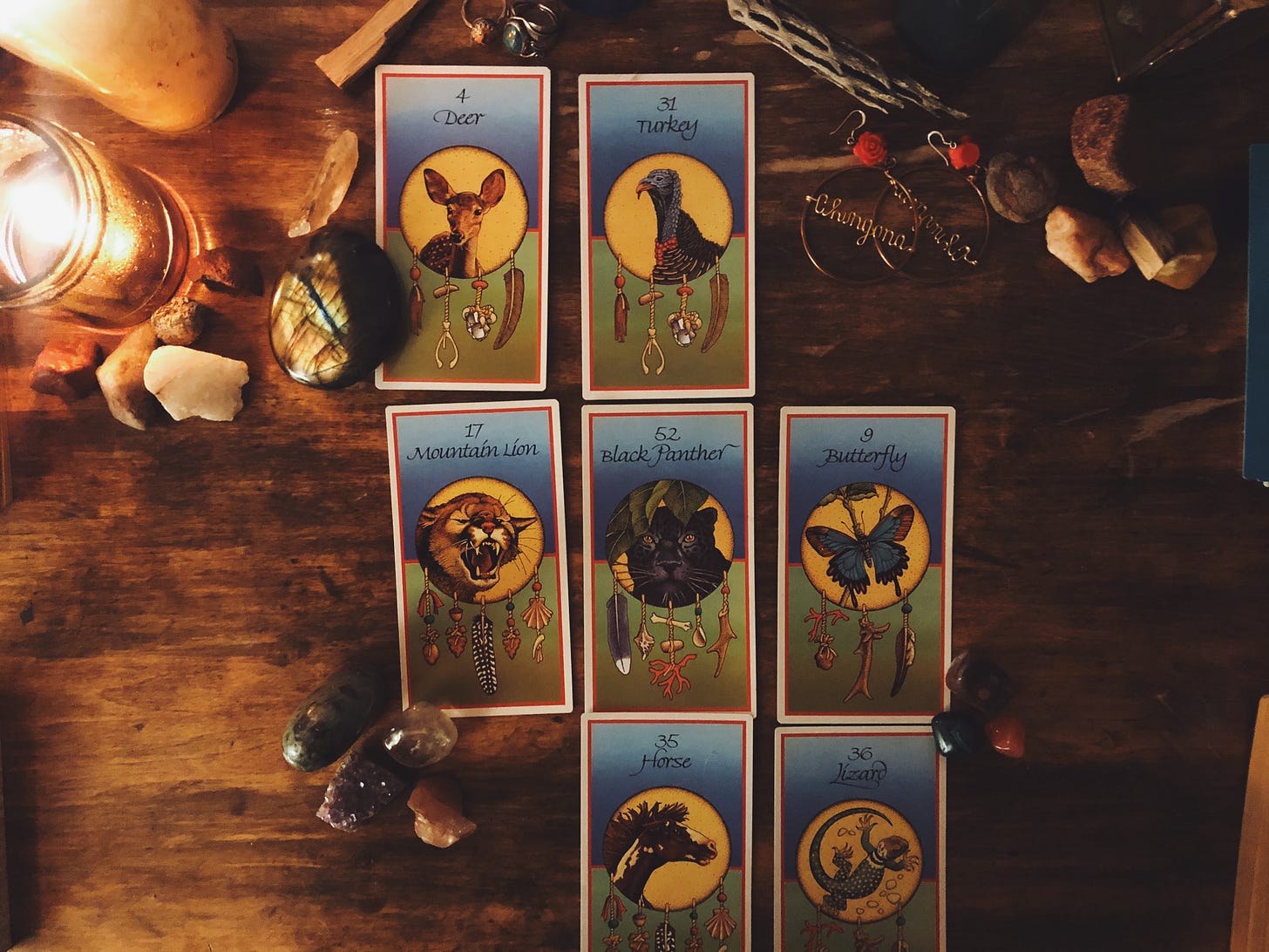 7 animal medicine oracle cards are set up in a Pathway spread, surrounded by candles and stones. From top left, the cards read: Deer, Turkey, Mountain Lion, Black Panther, Butterfly, Horse, and Lizard.