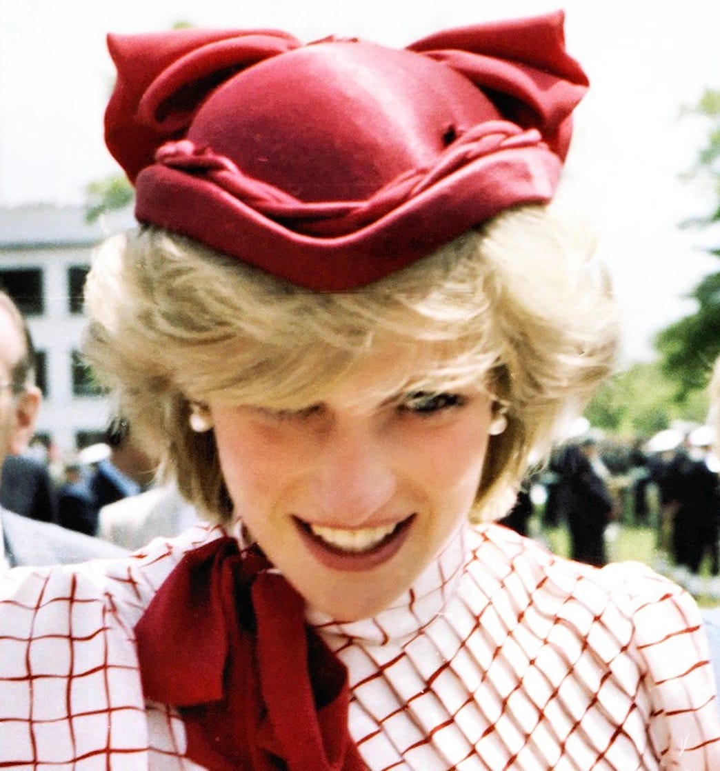Princess Diana is seen up close, face tilted downward, wearing a red and white checked blouse with a bright red sash and a bright red hat. Princess Diana is seen up close, face tilted downward, wearing a red and white checked blouse with a bright red sash and a bright red hat.