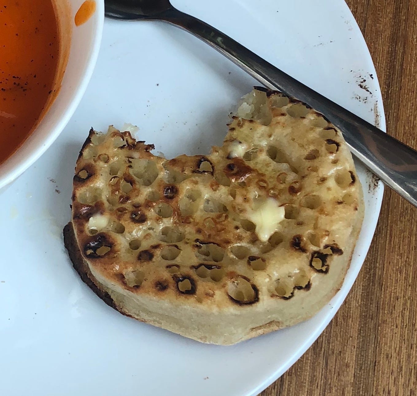 A closeup of a buttered toasted crumpet with a bite taken out of it, on the edge of a plate beside a red tomato soup. 