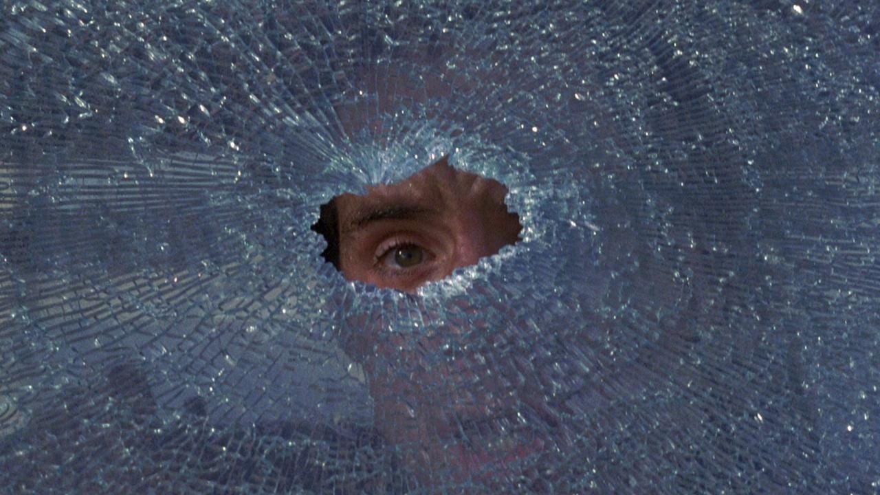 Film still from To Live and Die in L.A. A man's eye peeks through a hole in a shattered glass window.