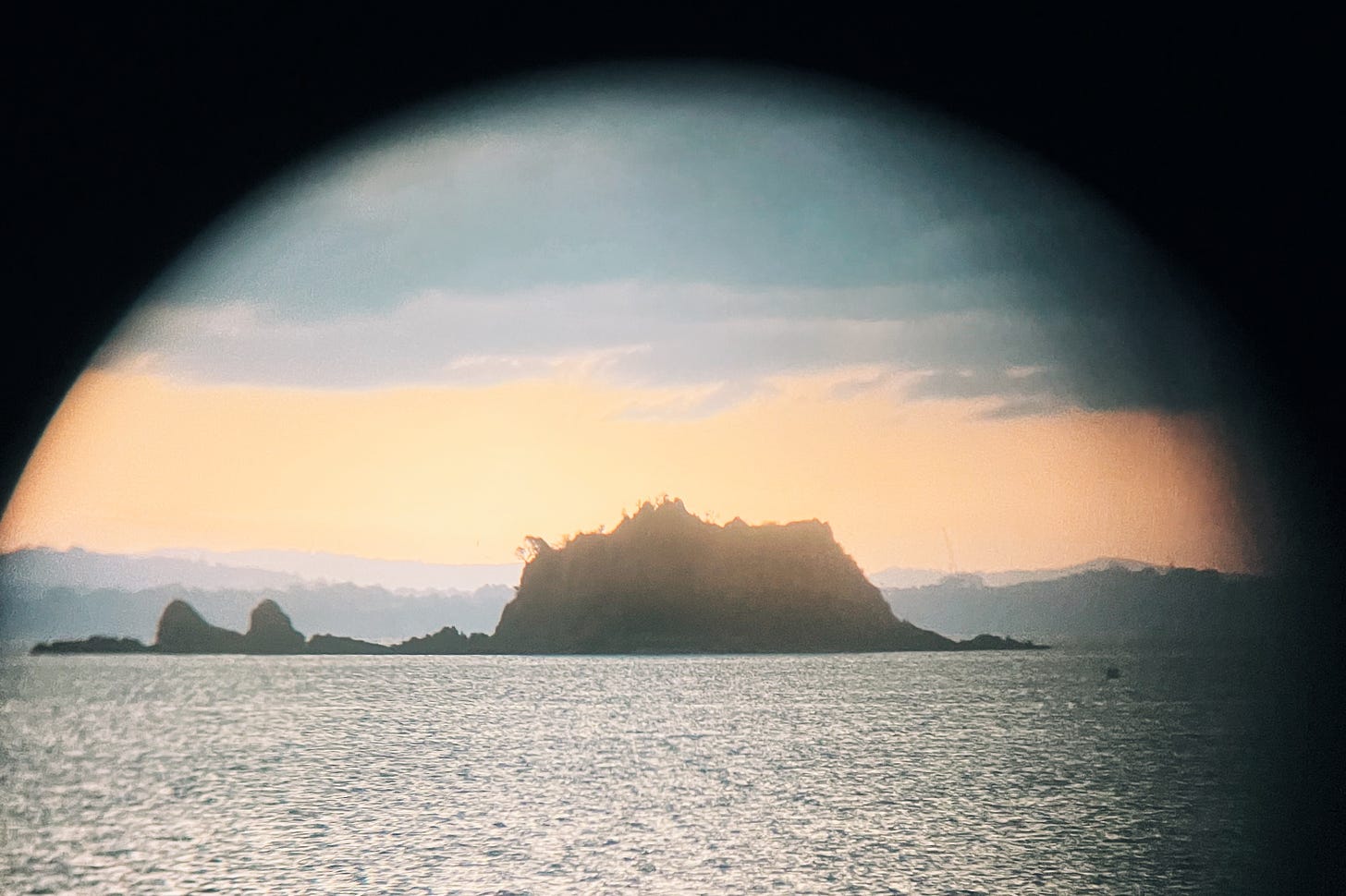 An island appears through the black border of a binoculars lens. The time is sundown and in the background is the sea and a layer of hills on a distant western shore