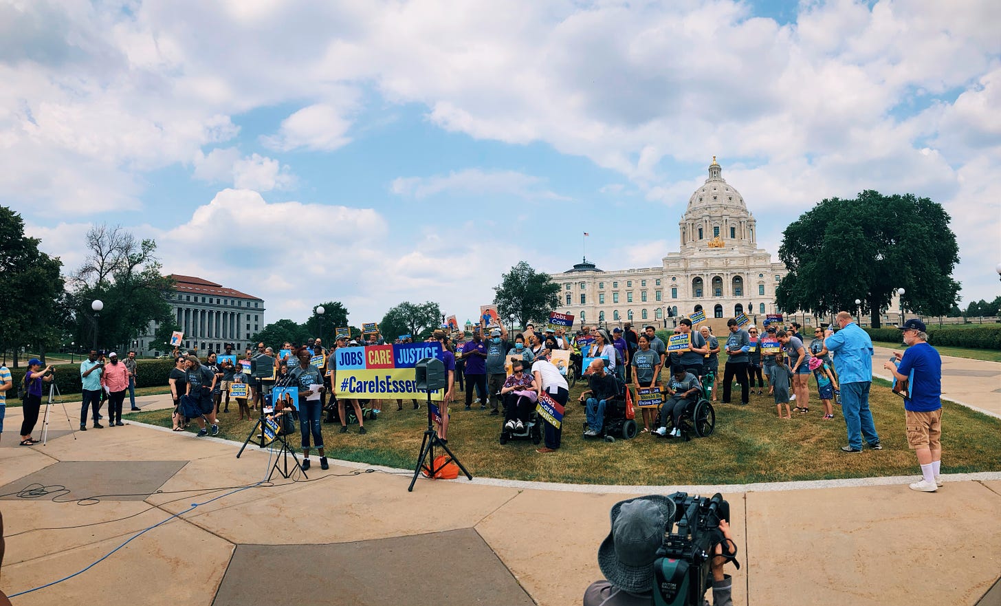 a crowd gathers on the front lawn of the state capitol building with signs saying "jobs care justice" as a newsperson points their camera at them