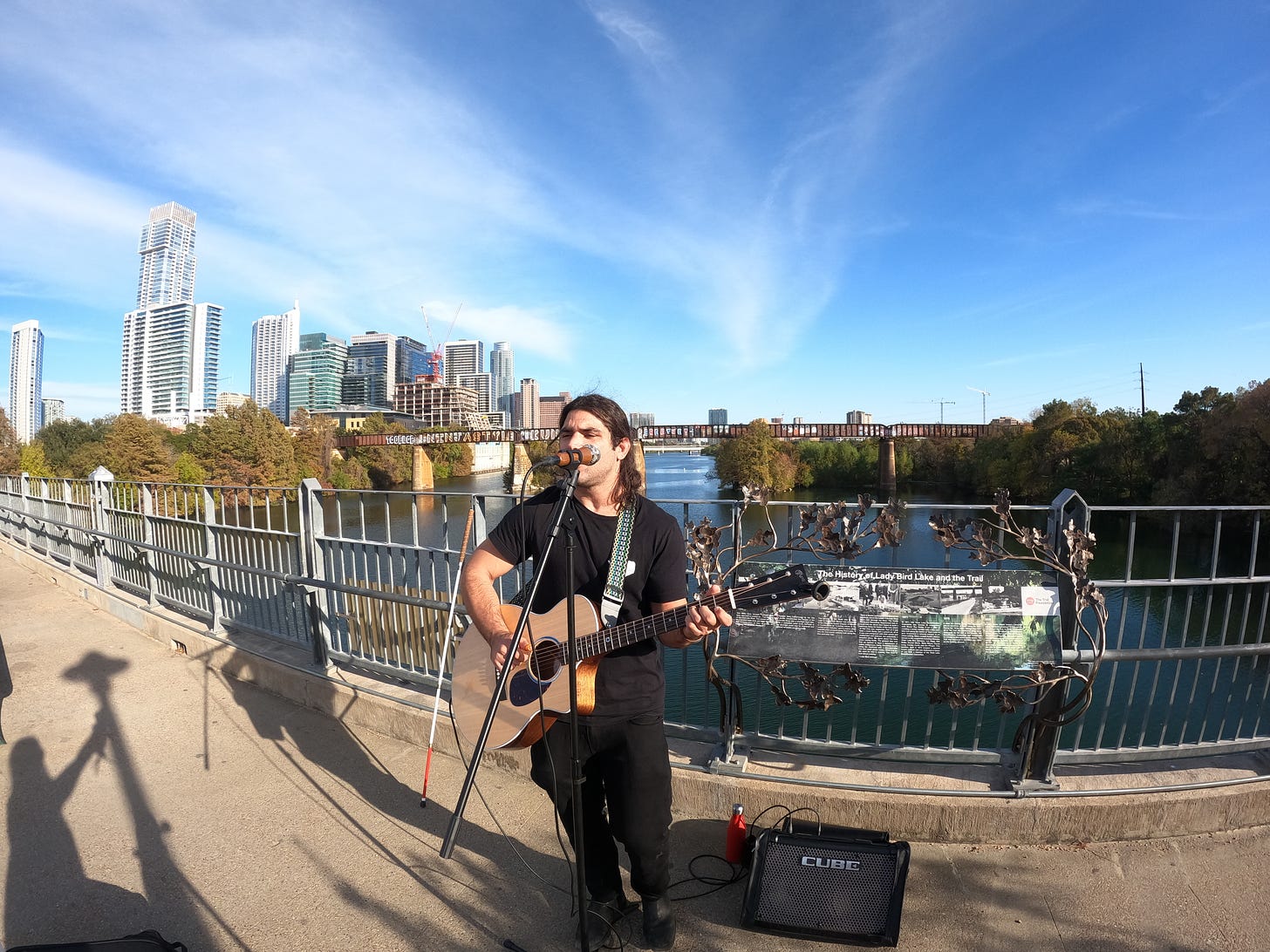 anthony standing on the bridge holding his guitar playing it with his white cane in the background and the city of austin texas in the background there is a river below him and the sky is blue and its nice and hot outside