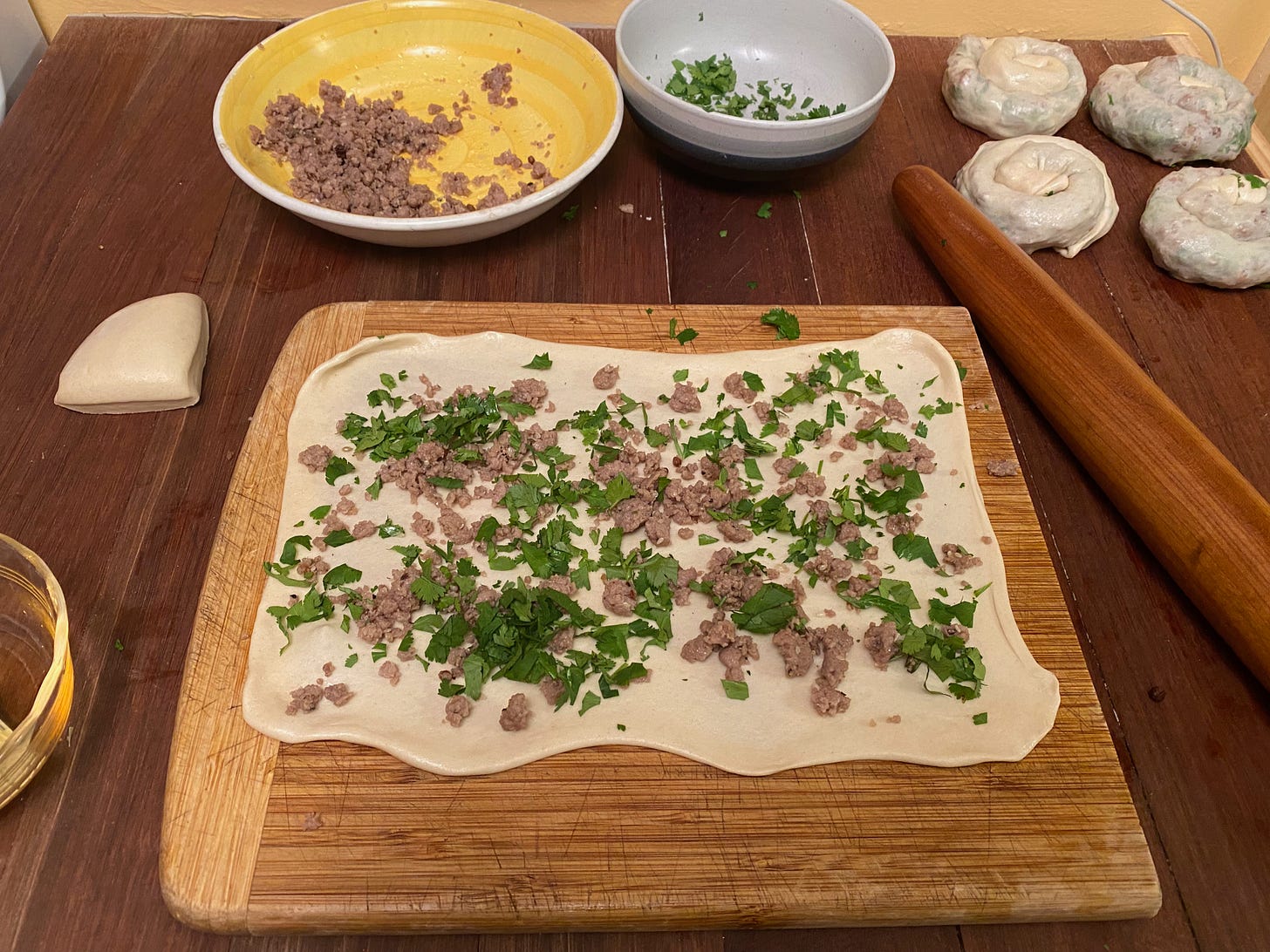 A large rectangle of rolled-out dough, scattered with pork and cilantro, sits on a cutting board on a wooden counter. Behind the cutting board is a bowl of pork, a bowl of cilantro leaves, a rolling pin, and four already-formed dough spirals.