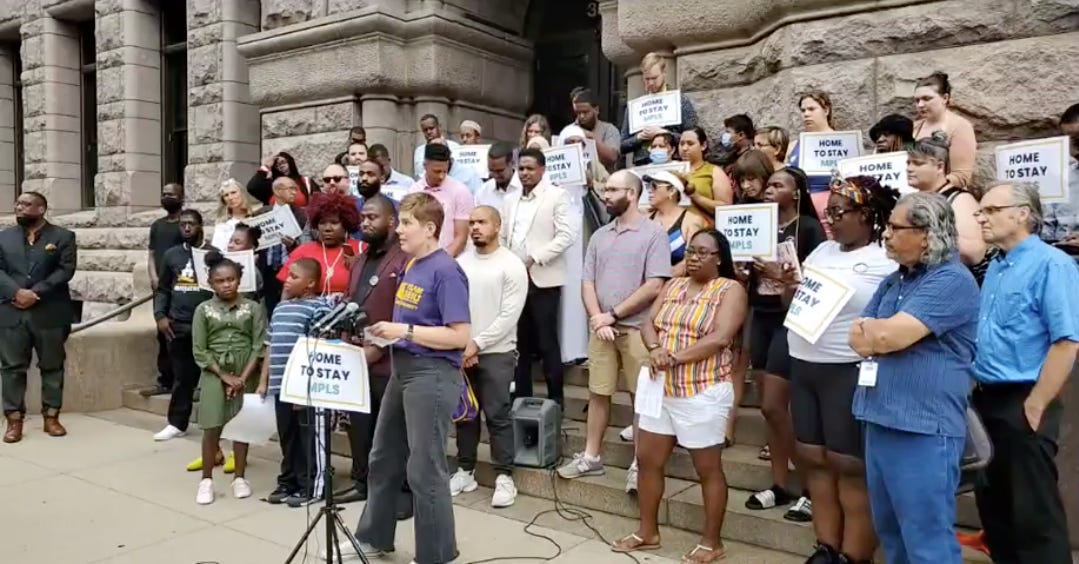 a large diverse group of people stand outside on the beige steps of city hall in front of a microphone set up