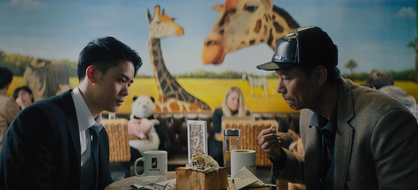 A father and grown son sit at a table and stare at a fancy cake between. The son on the left looks dejected, and the father on the right looks somewhat sullen but is raising his fork. Behind them people are dining and there's a giant giraffe mural on the wall.