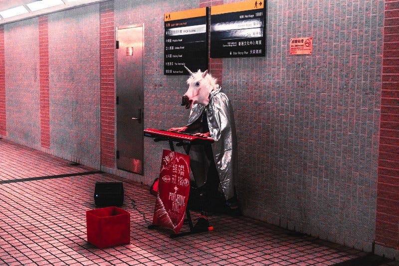 Person wearing a silver cape and unicorn mask playing a keyboard piano in a subway tunnel.