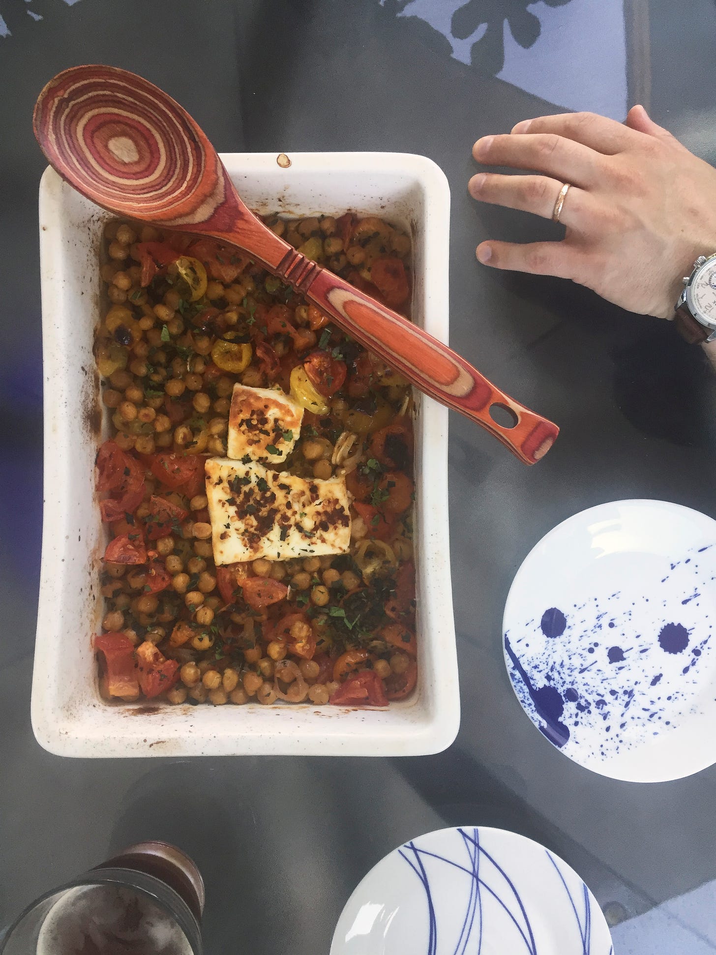 A white rectangular baking dish full of chickpeas and roasted tomatoes, with large pieces of feta covered in chili flakes at the centre. Herbs are sprinkled over the top and a wooden spoon sits crosswise at the top edge of the dish. Next to it on the table are two small plates, and Jeff's hand is just visible at the edge of the frame.