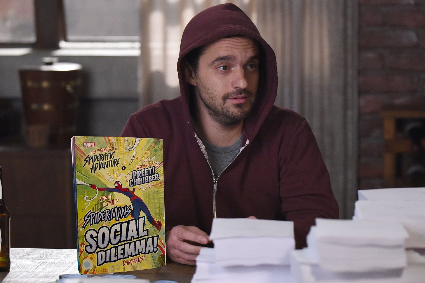 Jake Johnson in a hoodie sitting next to a photoshopped in copy of SPIDER-MAN'S SOCIAL DILEMMA