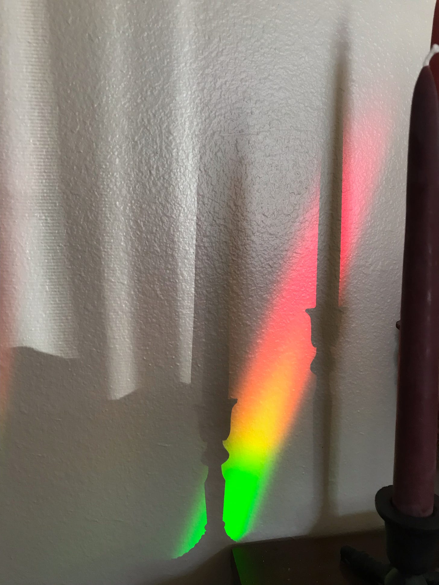 shadows of two taper candles and candlesticks on a white wall. a streak of red-yellow-green light shines on the wall.