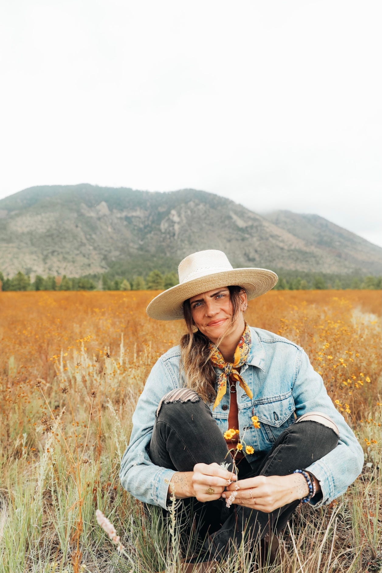 Ruthie Lindsey sitting in a field among the mountains, flowers in hand. 