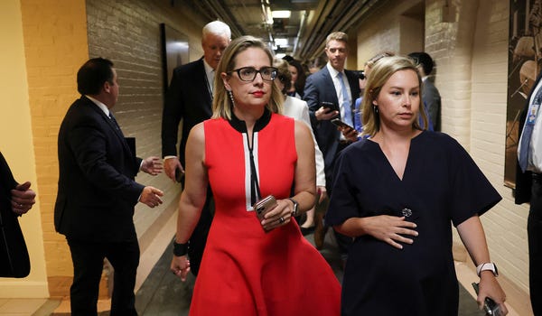 Senator Kyrsten Sinema walks down a crowded hallway, accompanied by another woman.