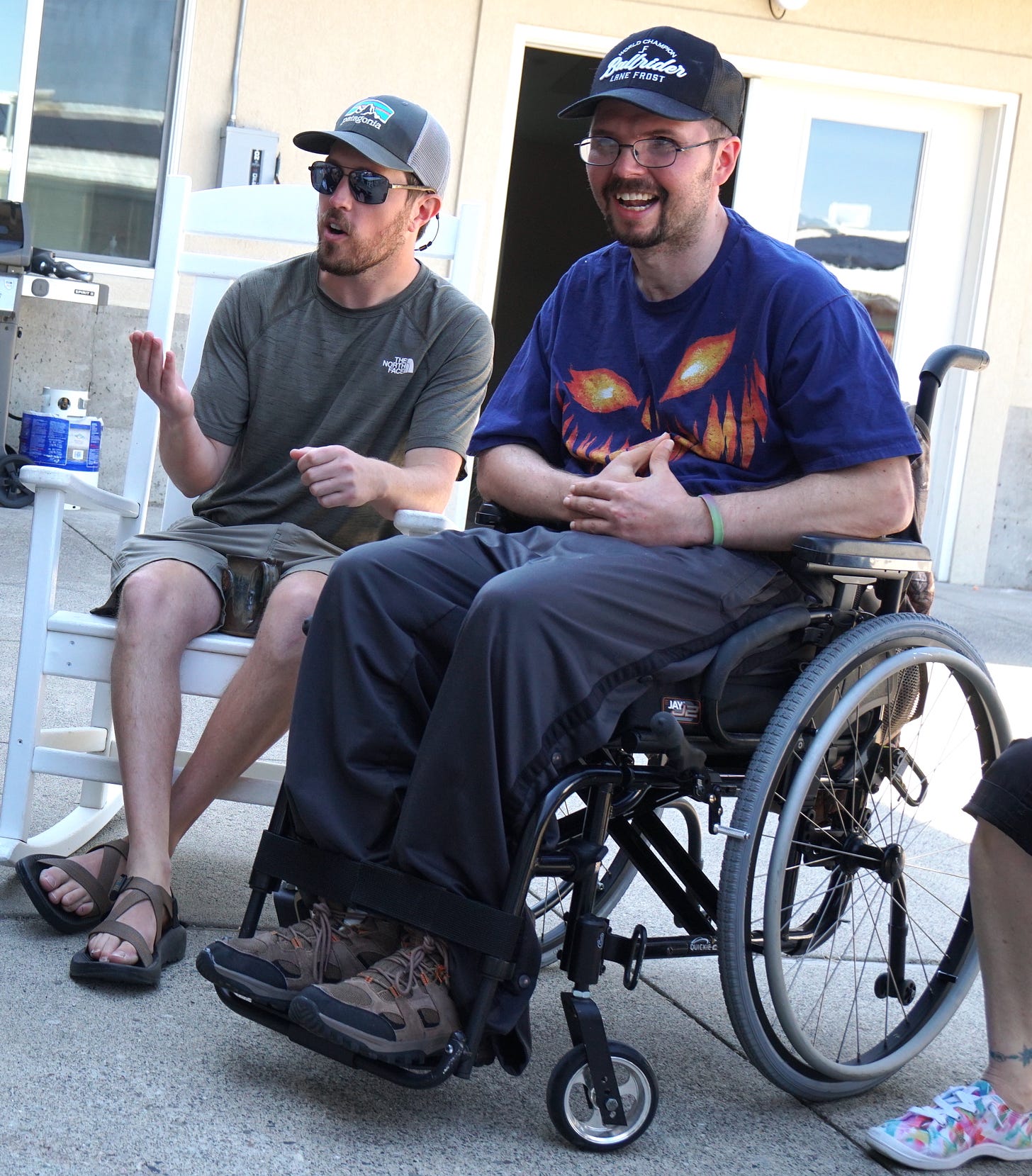 A man in a wheelchair with a goatee wearing a purple t-shirt, navy blue baseball cap and glasses smiles as another man with short facial hair sitting to his right wearing a gray t-shirt, cap and sunglasses gesticulates as he speaks to people outside the frame of the photo.