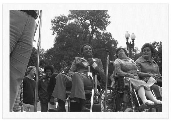 Brad Lomax, center, next to the activist Judy Heumann at a rally in 1977 at Lafayette Square in Washington. The photo is black and white. He is a Black man holding a mic, wearing a suit with wide lapels - very 70s - and seated in a manual wheelchair. Other disabled people are around him, on a stage and off.