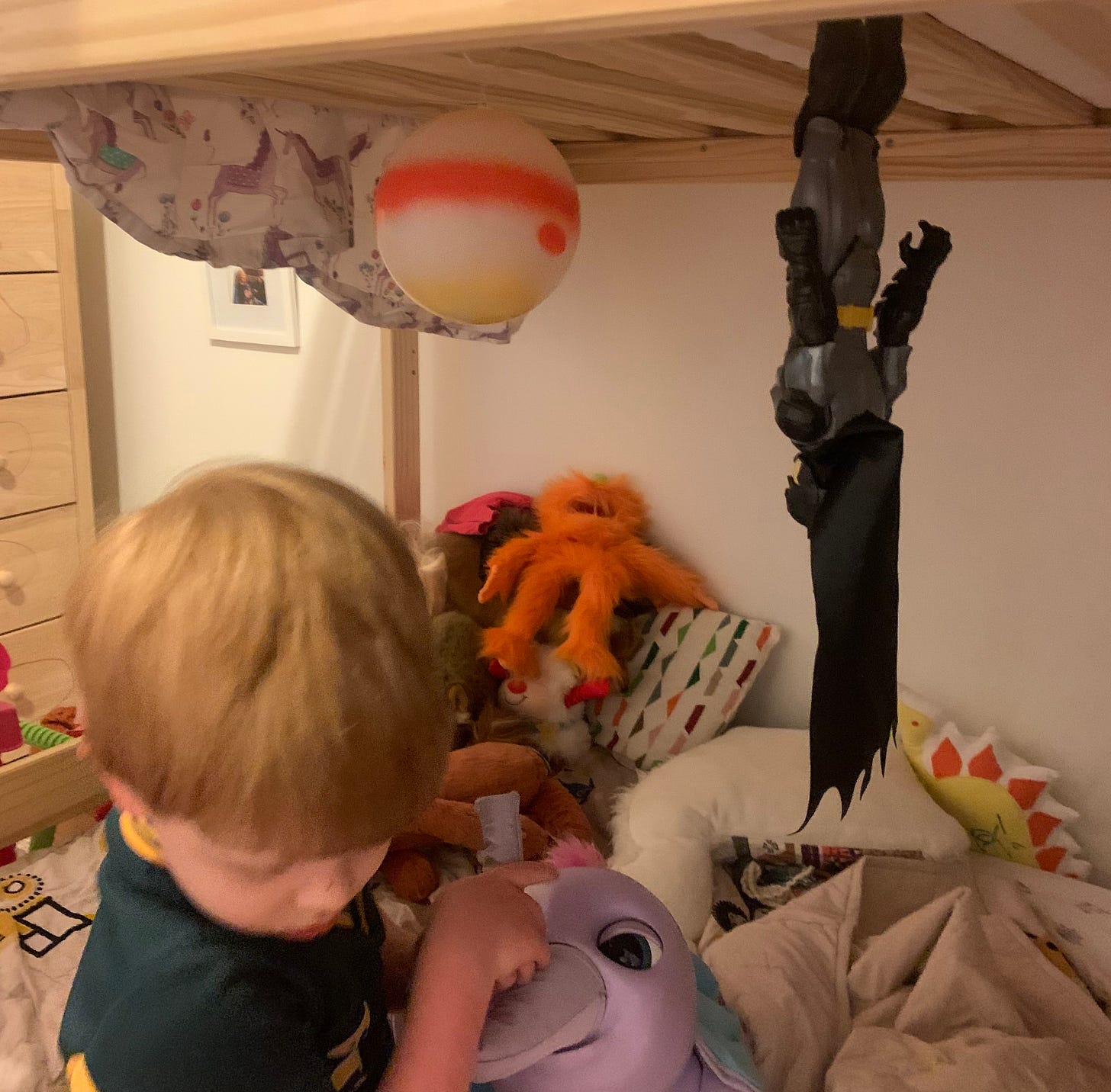Batman hangs upside down from a bunk bed, staring at a toy Jupiter, as a child plays