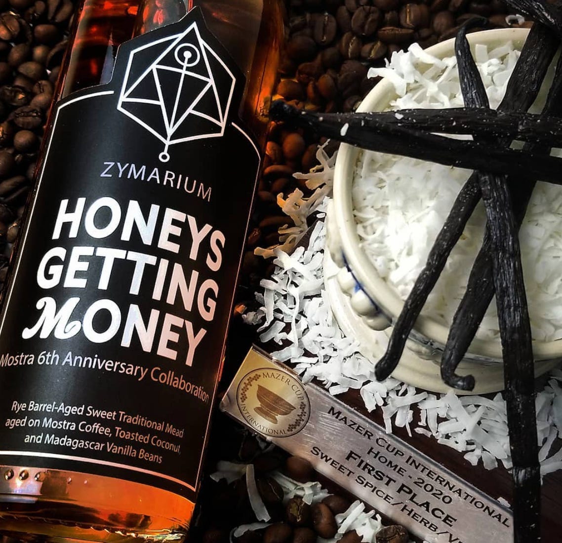 A bottle of honey mead resting on a bed of coffee beans, and to the right is a pile of shaved coconut and some vanilla beans.
