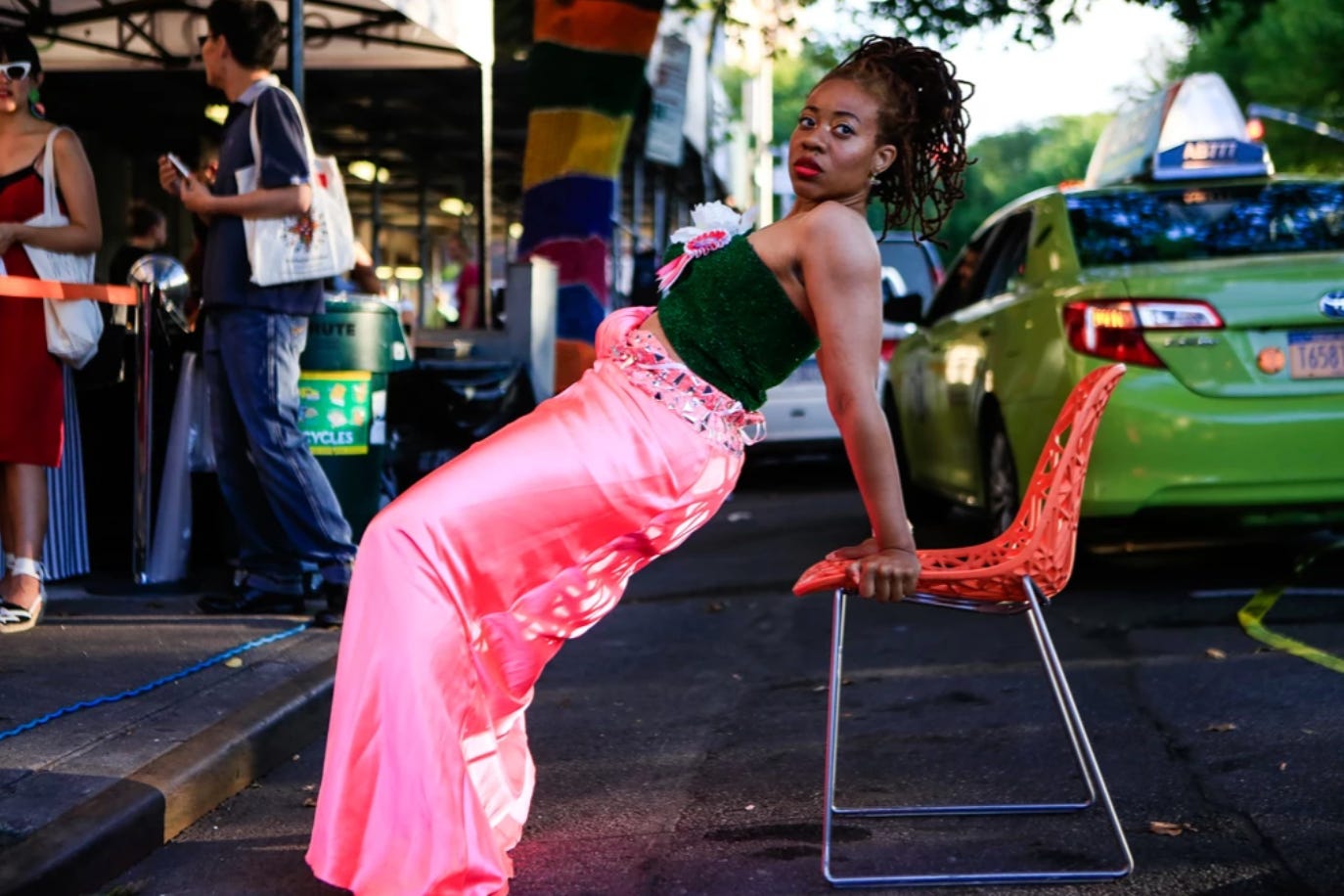 The artist wears a bright pink skirt and dark green top while looking forward. She is in the middle of performing bicep dips over a red chair on the middle of the street in New York. A green taxi drives off behind her on the road 