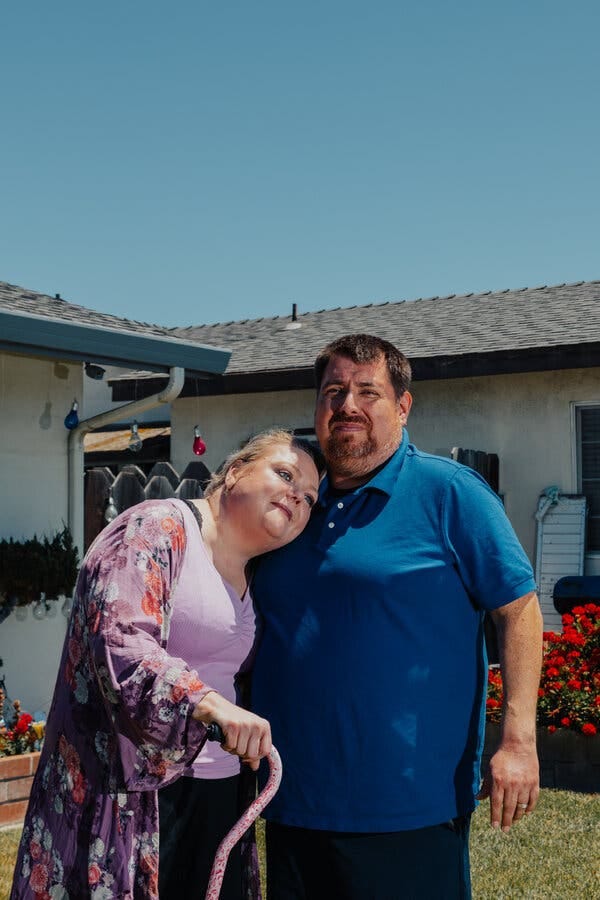 Two white people outside on a cloudless sunny day. One, wearing florals in purple and a matching cane, rests her head on the chest of her scruffy partner, in blue.