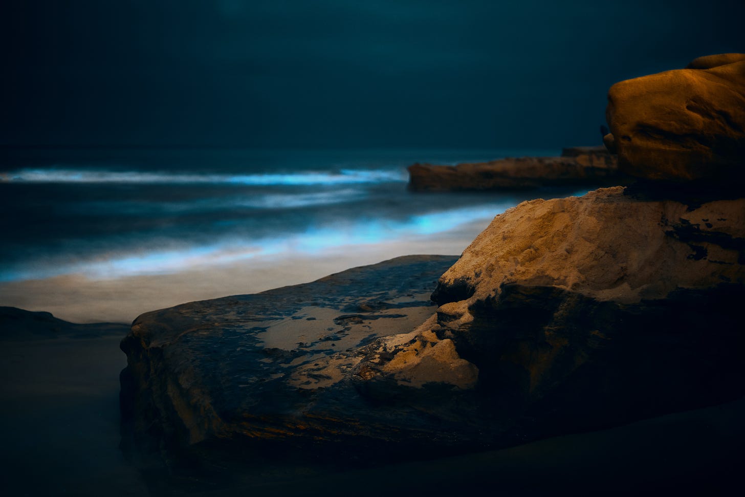A beach at night, with rocks in the foreground, a strip of sand behind them, and, in the hazy distance, a dark sea fringed with electric blue bioluminescence.