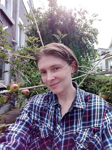 Portrait of Angeline B. Adams. She has short, light brown hair, blue eyes, and is wearing a blue plaid shirt. She is outdoors in front of an apple tree and some houses can be seen further in the background.