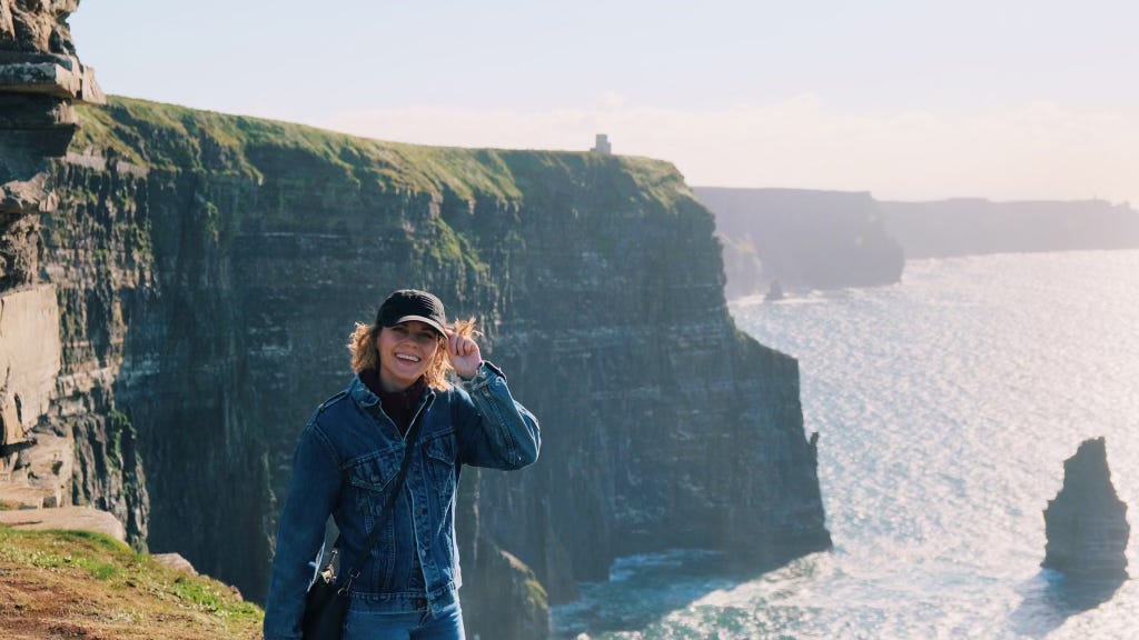 Leah, smiling and standing on the Cliffs of Moher on a sunny day.